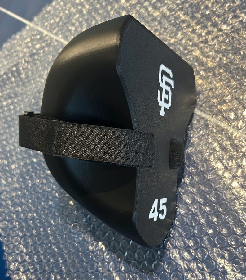 May include: A black foam knee pad featuring a white San Francisco Giants logo and the number 45 printed in white. The protective gear includes a black adjustable elastic strap with a hook and loop fastener for secure attachment during use.