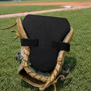 Pode incluir: Uma luva de beisebol de couro castanho desgastada com uma inserção de espuma preta fixada por duas tiras de velcro pretas. A luva apresenta atacadores visíveis ao longo das bordas e uma superfície texturizada na inserção preta.
