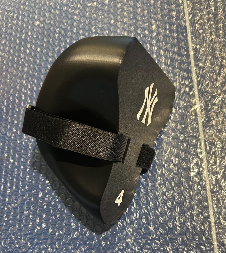 May include: A black foam knee pad featuring the white New York Yankees logo and the number 4 printed on the side. The protective gear includes a black adjustable fabric strap with a hook and loop fastener for secure attachment during use.