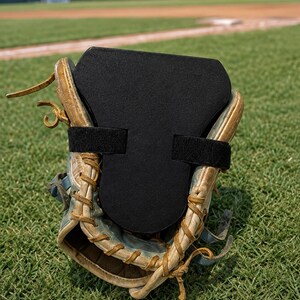 Puede incluir: Un guante de béisbol de cuero color canela desgastado con un inserto de espuma negra asegurado por dos correas de velcro negras. El guante presenta cordones visibles a lo largo de los bordes y una superficie texturizada en el inserto.