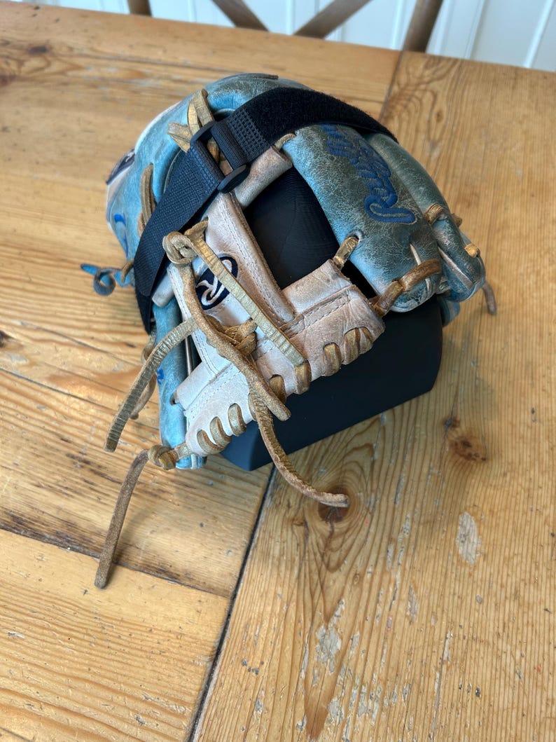 May include: A weathered light blue and white leather baseball glove featuring tan leather lacing, a black adjustable strap across the back, and the Rawlings brand logo embroidered in blue on the outer thumb section of the glove.