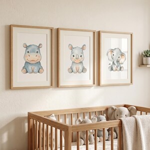 Pode incluir: Três impressões em aquarela emolduradas de animais bebês: um hipopótamo, um rinoceronte e um elefante. As impressões estão em molduras de madeira clara e são exibidas acima de um berço de madeira cheio de bichos de pelúcia e um cobertor. A obra de arte tem uma paleta de cores pastel.