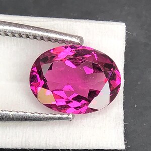 May include: A vibrant, oval-cut gemstone with a deep magenta-pink hue and intricate multifaceted surfaces that reflect light. The gemstone is held by a pair of silver-toned metal tweezers, showcasing its brilliant clarity and saturated color.