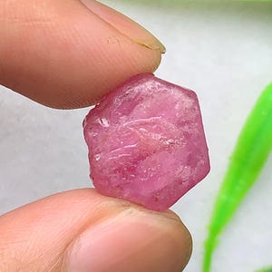 May include: A raw, hexagonal-shaped pink gemstone with a translucent, crystalline texture and natural surface inclusions. The stone displays varying shades of vibrant pink and magenta, featuring a rough, unpolished finish with distinct edges.