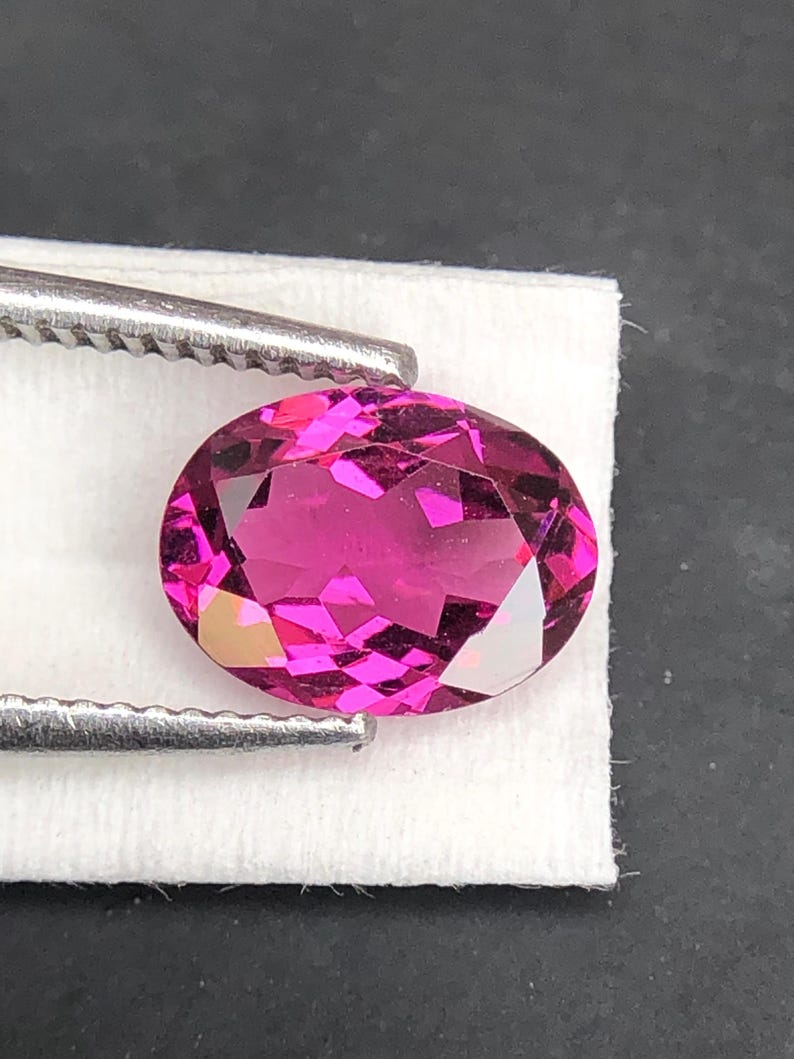 May include: A vibrant, oval-cut gemstone with a deep magenta-pink hue and intricate multifaceted surfaces that reflect light. The gemstone is held by a pair of silver-toned metal tweezers, showcasing its brilliant clarity and saturated color.