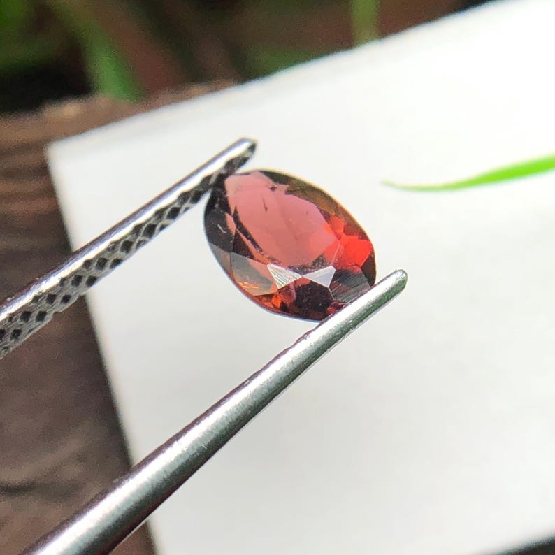 May include: A faceted, oval-shaped deep red gemstone held by a pair of silver-toned metal tweezers. The stone exhibits a translucent quality with visible internal light reflections and sharp, geometric cut edges across its polished surface.