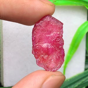 Può includere: Una gemma di rubino grezza e non lucidata con un colore rosso intenso e vibrante e una superficie cristallina strutturata. La pietra ha una forma irregolare e allungata con sfaccettature naturali visibili e inclusioni interne.