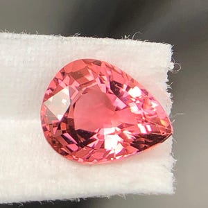 Può includere: Una pietra preziosa sfaccettata a forma di pera con una vibrante tonalità rosa-arancio. La pietra presenta molteplici sfaccettature triangolari e trapezoidali che riflettono la luce, creando un aspetto brillante e scintillante su una superficie bianca testurizzata.