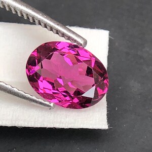 May include: An oval-cut, vibrant magenta gemstone with a faceted surface that reflects light. The gemstone is held by silver-toned metal tweezers against a small white rectangular paper base, showcasing its deep pink color and clarity.
