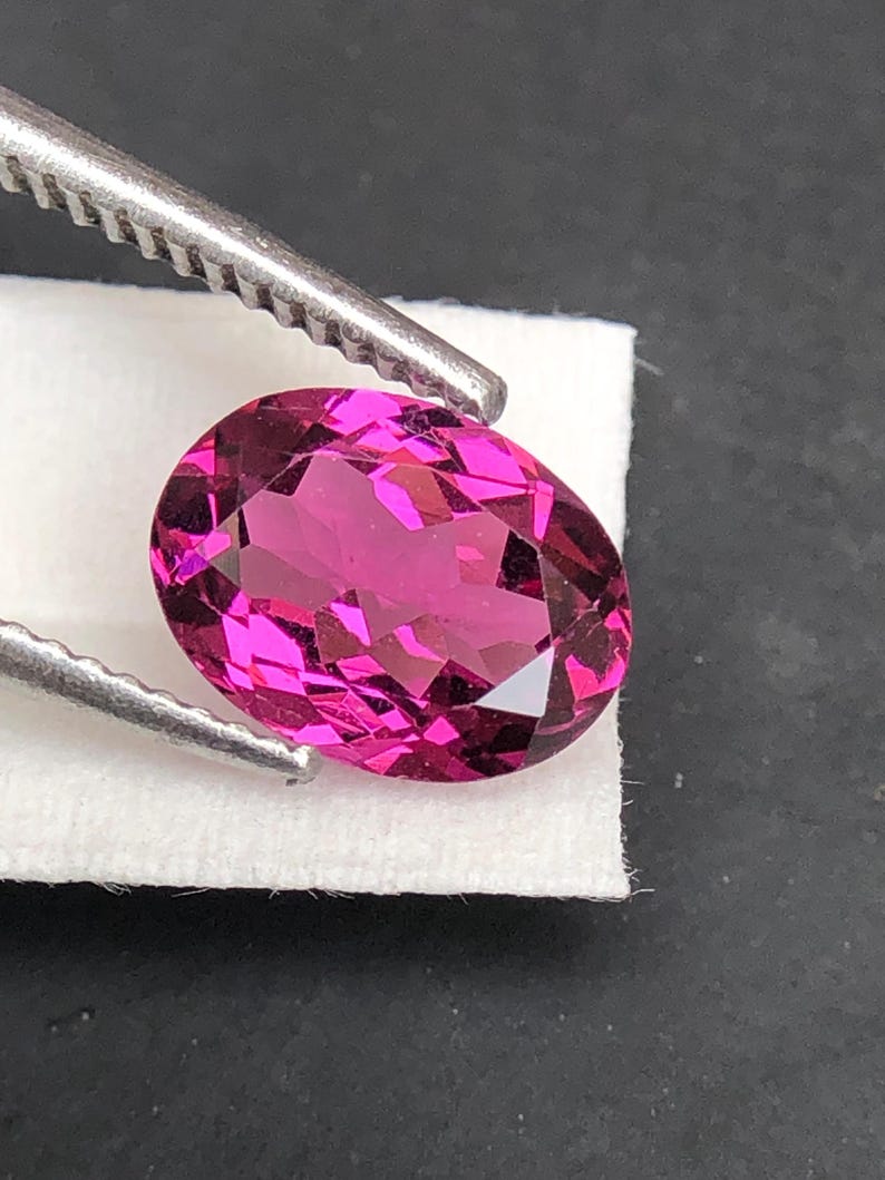 May include: An oval-cut, vibrant magenta gemstone with a faceted surface that reflects light. The gemstone is held by silver-toned metal tweezers against a small white rectangular paper base, showcasing its deep pink color and clarity.