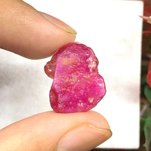 Può includere: Una pietra preziosa grezza e non lucidata con una vibrante tonalità rosso-rosata profonda. La pietra presenta una forma organica irregolare con una superficie cristallina testurizzata che cattura la luce, mostrando variazioni naturali di colore.