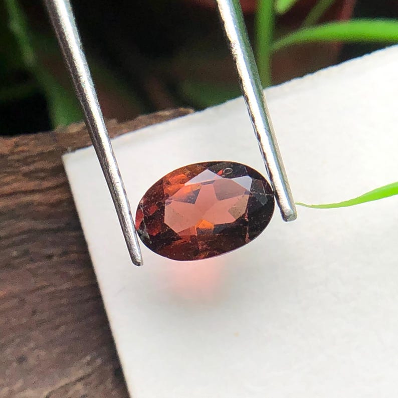 May include: An oval-shaped, faceted gemstone with a deep reddish-brown color held by a pair of silver-toned metal tweezers. The stone features a polished surface with multiple geometric facets that reflect light, showcasing its translucent quality.