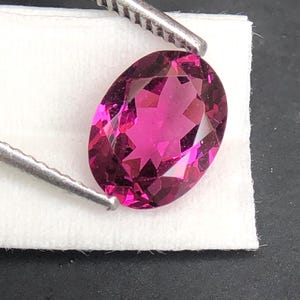 May include: A vibrant, oval-cut magenta gemstone with a faceted surface that reflects light. The stone is held by a pair of metal tweezers, showcasing its deep pink color and clear, transparent quality against a white textured background.