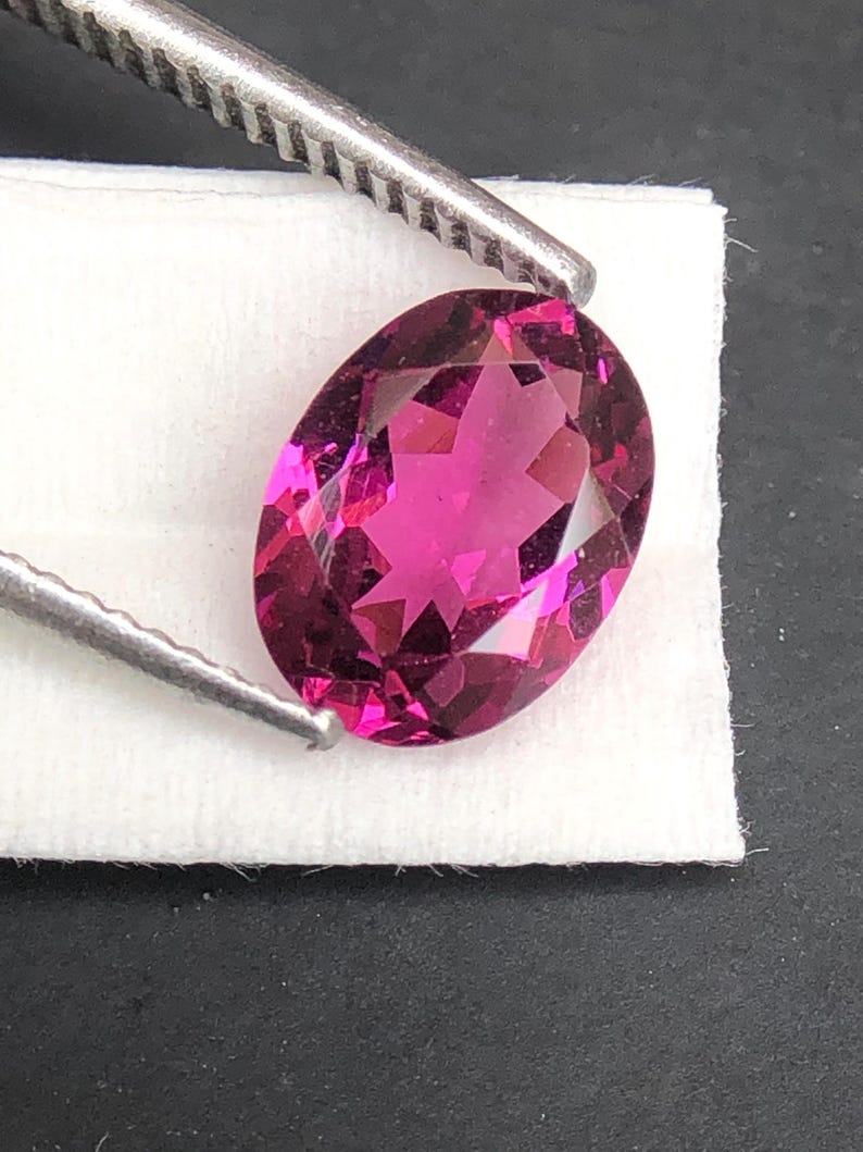 May include: A vibrant, oval-cut magenta gemstone with a faceted surface that reflects light. The stone is held by a pair of metal tweezers, showcasing its deep pink color and clear, transparent quality against a white textured background.