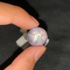 May include: A round, cabochon-cut star sapphire gemstone with a light lavender-purple hue. The stone features a distinct six-rayed star effect centered on its surface, accompanied by small, dark, natural inclusions scattered across the polished surface.