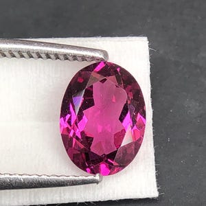 May include: An oval-cut, deep magenta gemstone with multiple facets that reflect light. The stone is held by silver-toned metal tweezers against a small, white rectangular paper backing, showcasing its vibrant color and clear, polished surface.