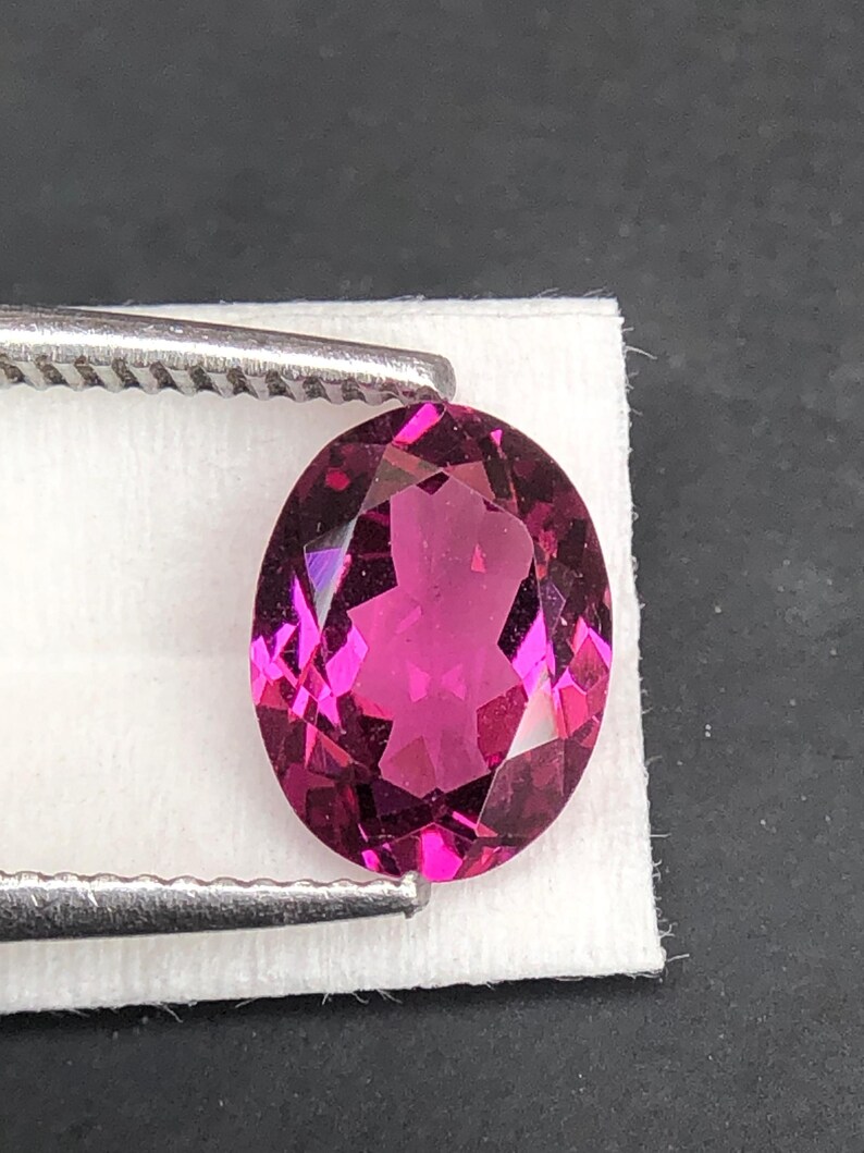 May include: An oval-cut, deep magenta gemstone with multiple facets that reflect light. The stone is held by silver-toned metal tweezers against a small, white rectangular paper backing, showcasing its vibrant color and clear, polished surface.