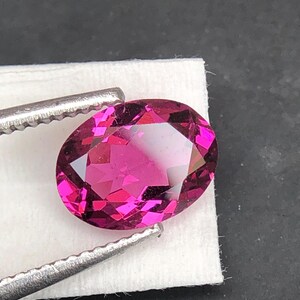 May include: An oval-cut, vibrant magenta gemstone with a faceted surface that reflects light. The gemstone is held by a pair of silver-toned metal tweezers, showcasing its deep pink color, clarity, and precise geometric cut against a white base.