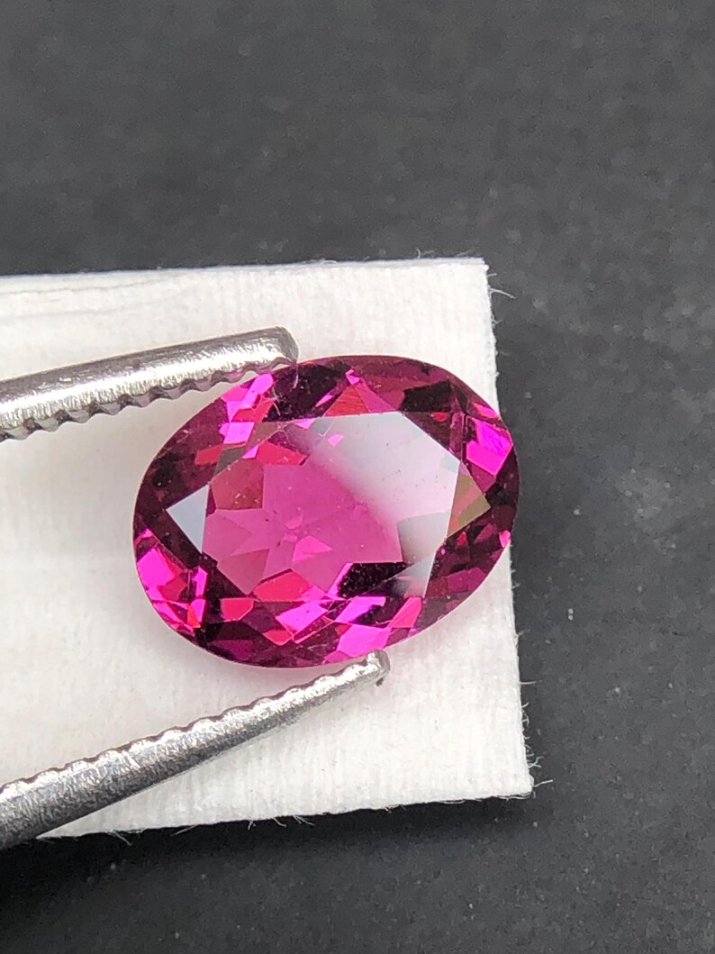 May include: An oval-cut, vibrant magenta gemstone with a faceted surface that reflects light. The gemstone is held by a pair of silver-toned metal tweezers, showcasing its deep pink color, clarity, and precise geometric cut against a white base.