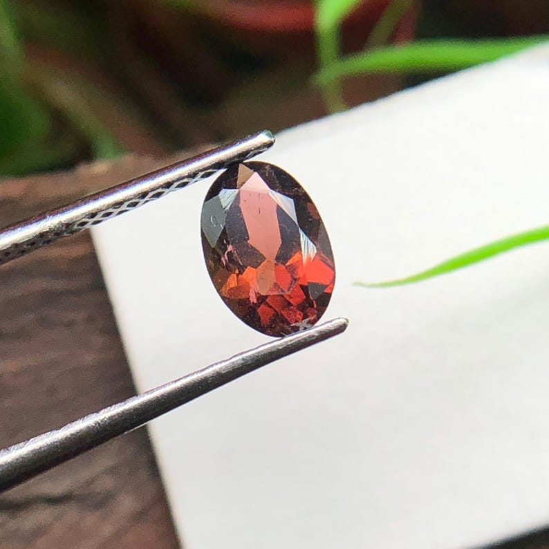 May include: A faceted oval-shaped deep red gemstone held by metal tweezers. The stone exhibits a translucent quality with visible internal light reflections and sharp geometric cuts across its surface, showcasing a rich, dark crimson color profile.