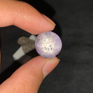 May include: A round, cabochon-cut lavender gemstone featuring a distinct six-rayed star effect centered on its surface. The stone displays a translucent, pale purple hue with subtle dark inclusions and a polished, smooth, domed top surface.