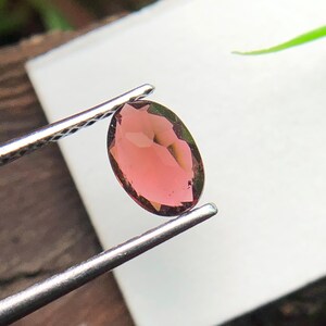 May include: An oval-shaped, faceted gemstone with a translucent pinkish-red hue held by a pair of silver-toned metal tweezers. The stone features multiple geometric facets that reflect light, creating a polished and clear appearance throughout.