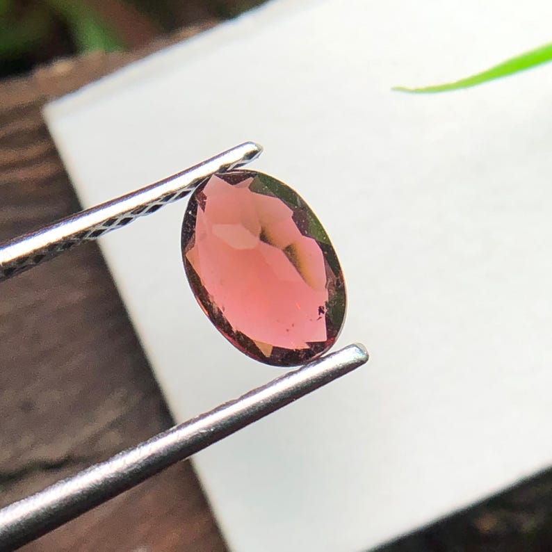 May include: An oval-shaped, faceted gemstone with a translucent pinkish-red hue held by a pair of silver-toned metal tweezers. The stone features multiple geometric facets that reflect light, creating a polished and clear appearance throughout.
