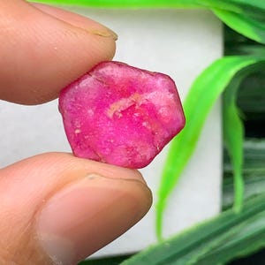 Può includere: Una pietra preziosa grezza e non tagliata di un colore rosa acceso e saturo. La pietra ha una forma irregolare, leggermente esagonale, con una superficie ruvida e leggermente traslucida che mostra inclusioni naturali e sfaccettature.
