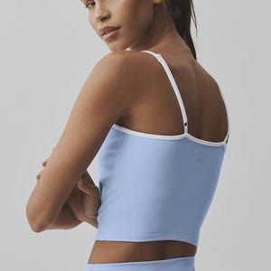 May include: Pale blue crop top with white trim and straps. The athletic top has a scoop neck and is paired with matching pale blue leggings. The model is wearing a ponytail and looking over their shoulder.