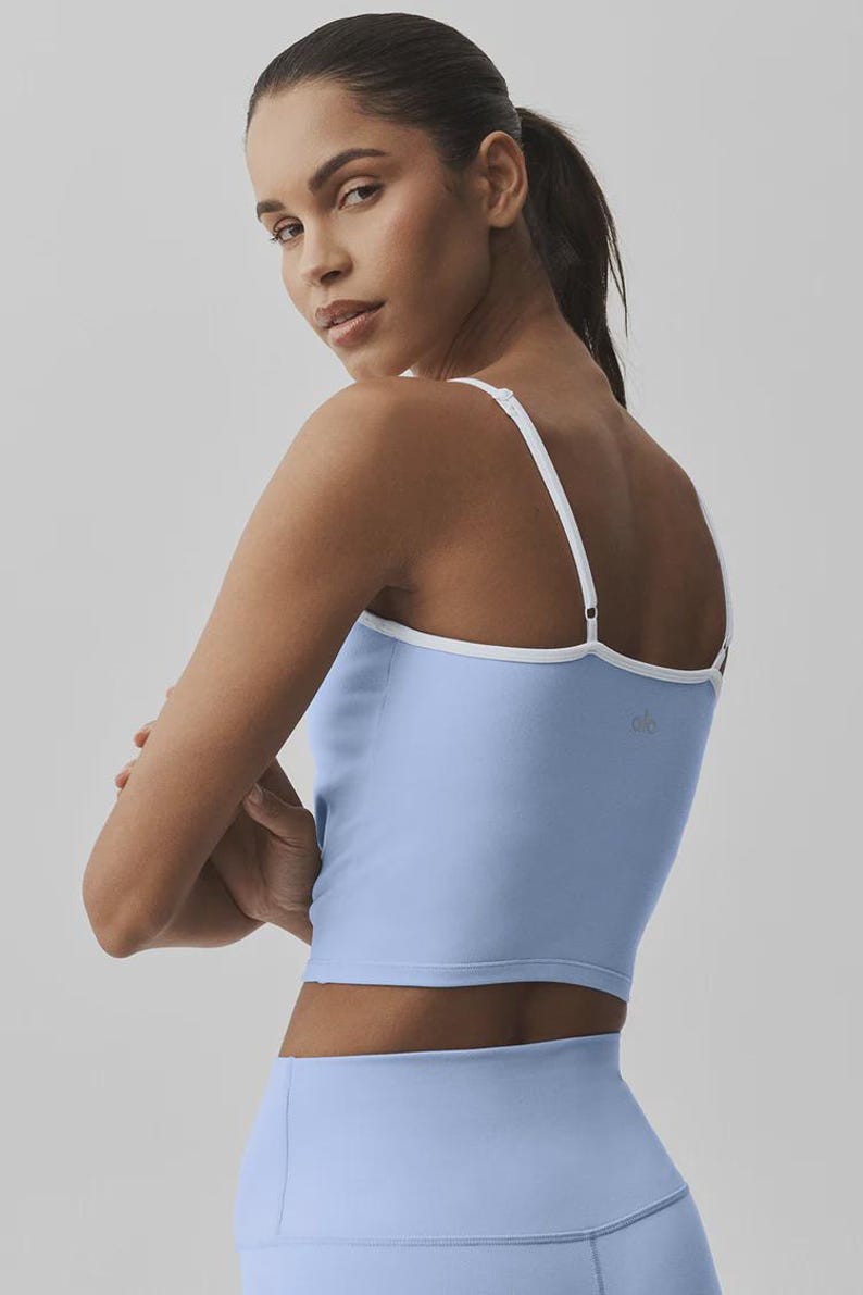 May include: Pale blue crop top with white trim and straps. The athletic top has a scoop neck and is paired with matching pale blue leggings. The model is wearing a ponytail and looking over their shoulder.
