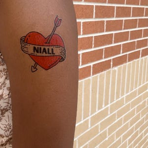 May include: A temporary tattoo featuring a bright red heart pierced by an arrow. A beige banner wraps around the center of the heart with the name NIALL printed in bold black capital letters. The design has a black outline and a classic aesthetic.