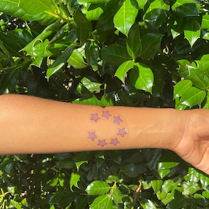 May include: A temporary tattoo applied to the skin featuring a circular arrangement of seven small, purple, five-pointed stars. The design is printed on a clear, circular adhesive backing that is centered on the forearm.
