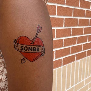 May include: A temporary tattoo featuring a red heart pierced by an arrow. A beige banner wraps around the heart with the text SOMBR printed in black capital letters. The design uses bold black outlines and is applied to the skin of an upper arm.