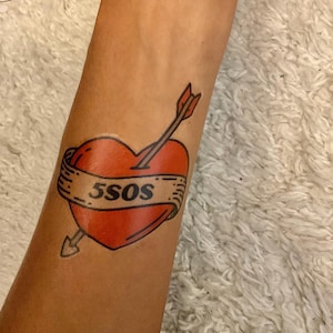 May include: A temporary tattoo featuring a vibrant red heart pierced by a dark arrow. A beige banner wraps around the center of the heart, displaying the black text 5SOS in a bold, sans-serif font. The design uses clean black outlines.