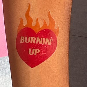 May include: A temporary tattoo featuring a solid red heart shape topped with orange flames. The text "BURNIN UP" is printed in a bold, light-colored sans-serif font across the center of the heart, creating a clear and graphic design.