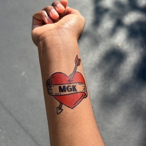 May include: A temporary tattoo on a forearm featuring a red heart pierced by an arrow. A beige banner wraps around the heart with the letters MGK printed in bold black text. The design uses thick black outlines and classic tattoo-style shading.