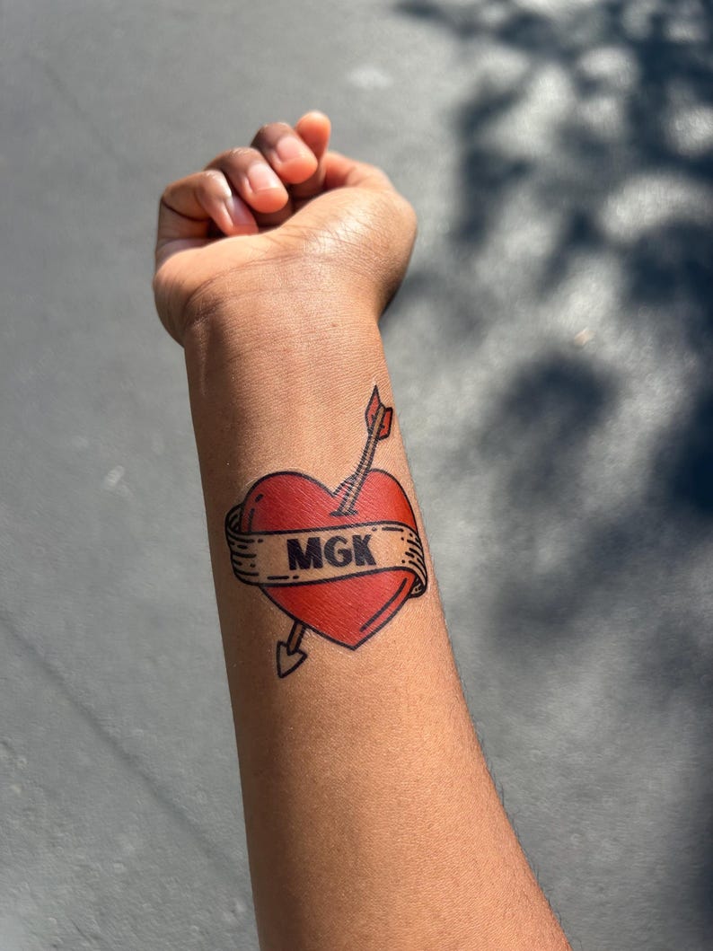 May include: A temporary tattoo on a forearm featuring a red heart pierced by an arrow. A beige banner wraps around the heart with the letters MGK printed in bold black text. The design uses thick black outlines and classic tattoo-style shading.