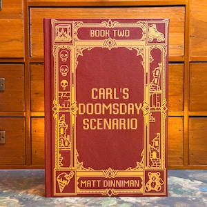Puede incluir: Un libro de tapa dura titulado Carl's Doomsday Scenario Book Two de Matt Dinniman. La cubierta es de color rojo intenso con detalles en lámina dorada, con iconos pixelados de calaveras, un patín, un helado, un oso de peluche y edificios en ruinas.