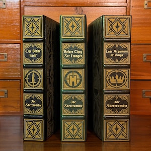 May include: Three hardback books with dark green spines and gold embossed detailing. The spines feature geometric patterns, sword, tower, and crown icons, and the titles The Blade Itself, Before They Are Hanged, and Last Argument of Kings.