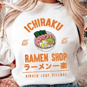 May include: A white crewneck sweatshirt featuring a graphic of a bowl of ramen with the text ICHIRAKU RAMEN SHOP, Japanese characters, and HIDDEN LEAF VILLAGE. The design includes orange lettering and two spiral symbols on either side of the bowl.