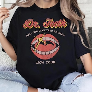 Könnte beinhalten: Ein schwarzes Kurzarm-T-Shirt mit einer Grafik eines offenen Mundes mit Zähnen und einem gelben Kreuz. Der Text lautet Dr. Teeth and the Electric Mayhem und 1976 Tour in einer Retro-Schriftart. Das Design hat ein abgenutztes Aussehen.