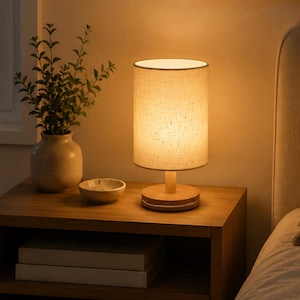 May include: A cylindrical table lamp with a light-colored fabric shade and a circular wooden base. The lamp emits a warm, soft glow, illuminating the surrounding area. It features a simple, clean design with a natural wood finish on the base.