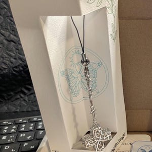 May include: A silver-toned metal cross charm with an intricate filigree cutout design, suspended from a black cord. The charm is attached to a white display card featuring a butterfly illustration and the text IvoryIvy Imaginings.