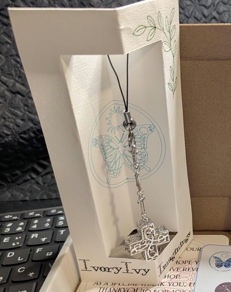 May include: A silver-toned metal cross charm with an intricate filigree cutout design, suspended from a black cord. The charm is attached to a white display card featuring a butterfly illustration and the text IvoryIvy Imaginings.
