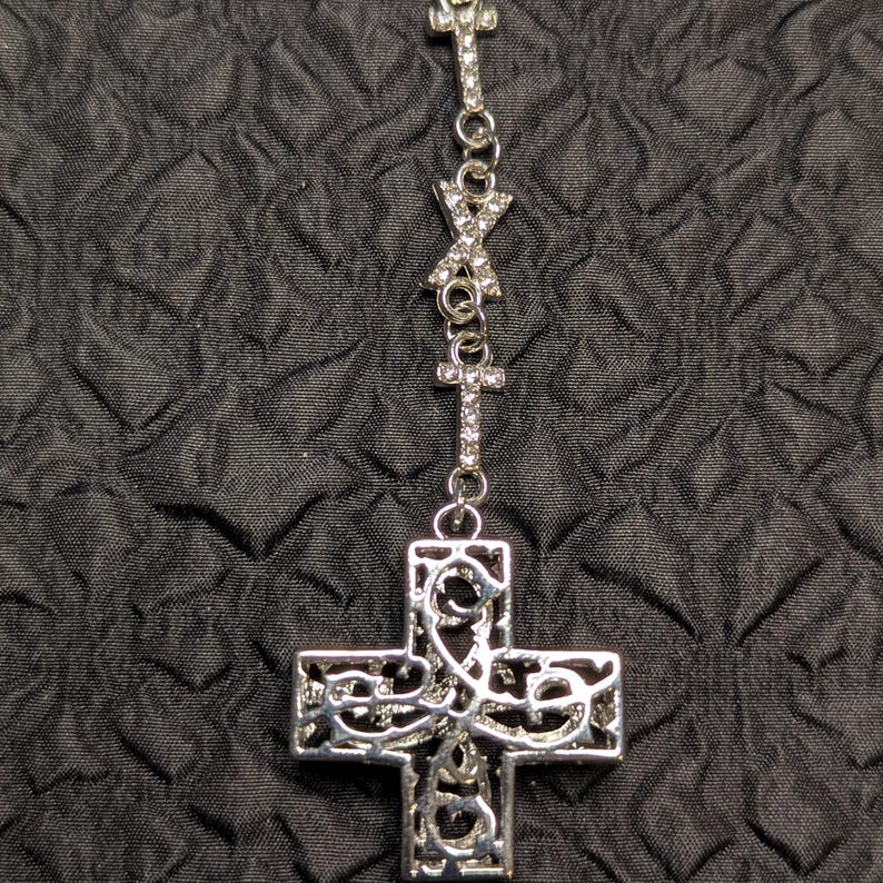 May include: A silver-toned necklace featuring a large, ornate cross pendant with intricate filigree cutouts. The chain is adorned with smaller cross-shaped links and an X-shaped link, all encrusted with small, clear, sparkling rhinestones.