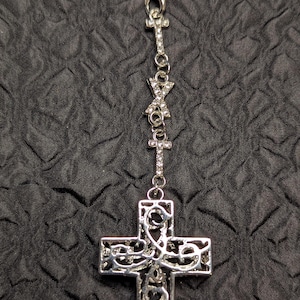 May include: A silver-toned metal charm featuring a filigree cross pendant suspended from a chain adorned with two smaller crystal-encrusted crosses and a central crystal-studded X-shaped connector, all attached to a black cord loop.