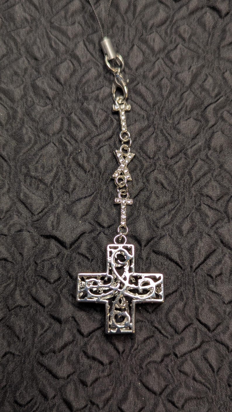 May include: A silver-toned metal charm featuring a filigree cross pendant suspended from a chain adorned with two smaller crystal-encrusted crosses and a central crystal-studded X-shaped connector, all attached to a black cord loop.