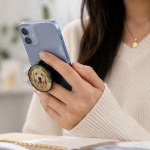 Peut inclure: Un smartphone bleu clair doté d'un support de téléphone circulaire rétractable fixé au dos. Le support présente une impression photographique d'un golden retriever la gueule ouverte, affichant une expression joyeuse sur un fond vert doux.