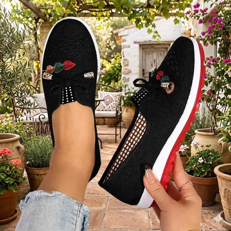 May include: Black slip-on shoes with a decorative floral design. The shoes have a white sole with a red stripe, and a black bow detail. The shoes are decorated with red, green, and gold accents.