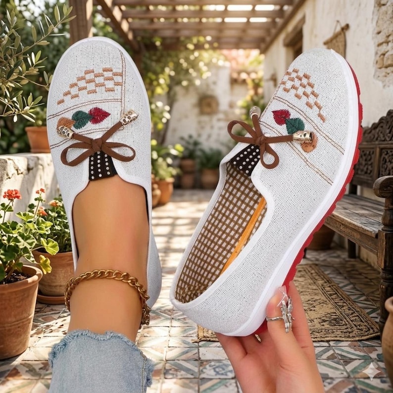 May include: White slip-on shoes with embroidered details. The shoes feature a brown bow, red soles, and a patterned design on the top. The interior has a brown and white checkered pattern. The shoes are designed for casual wear.